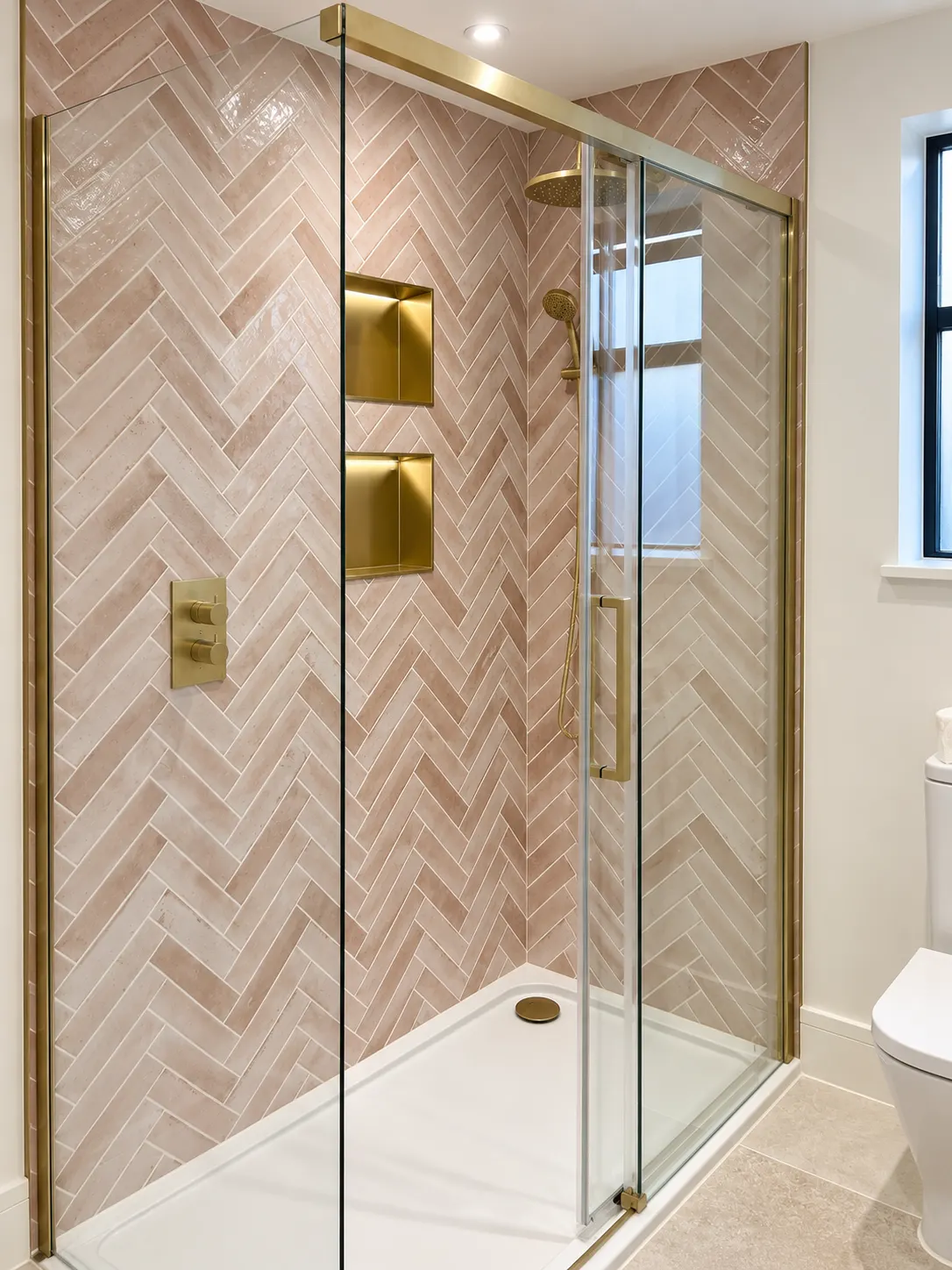 Pink herringbone tiled shower with brushed brass valve, lit niches and a sliding glass door